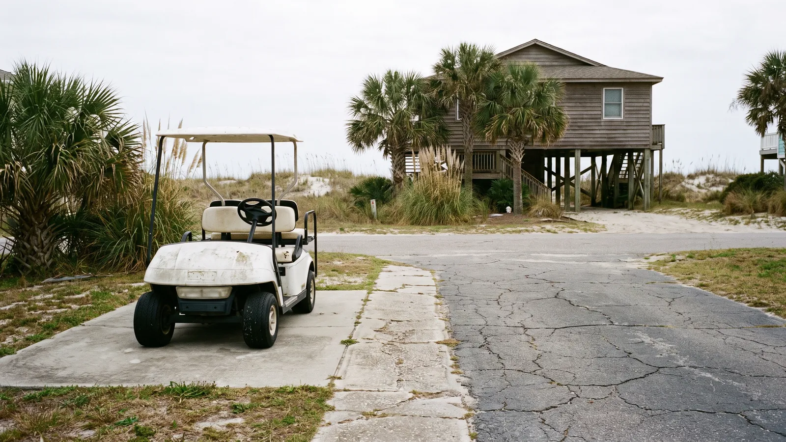 Concrete golf cart pad and narrow path beside a raised beach house with a golf cart parked on it, dune grass behind